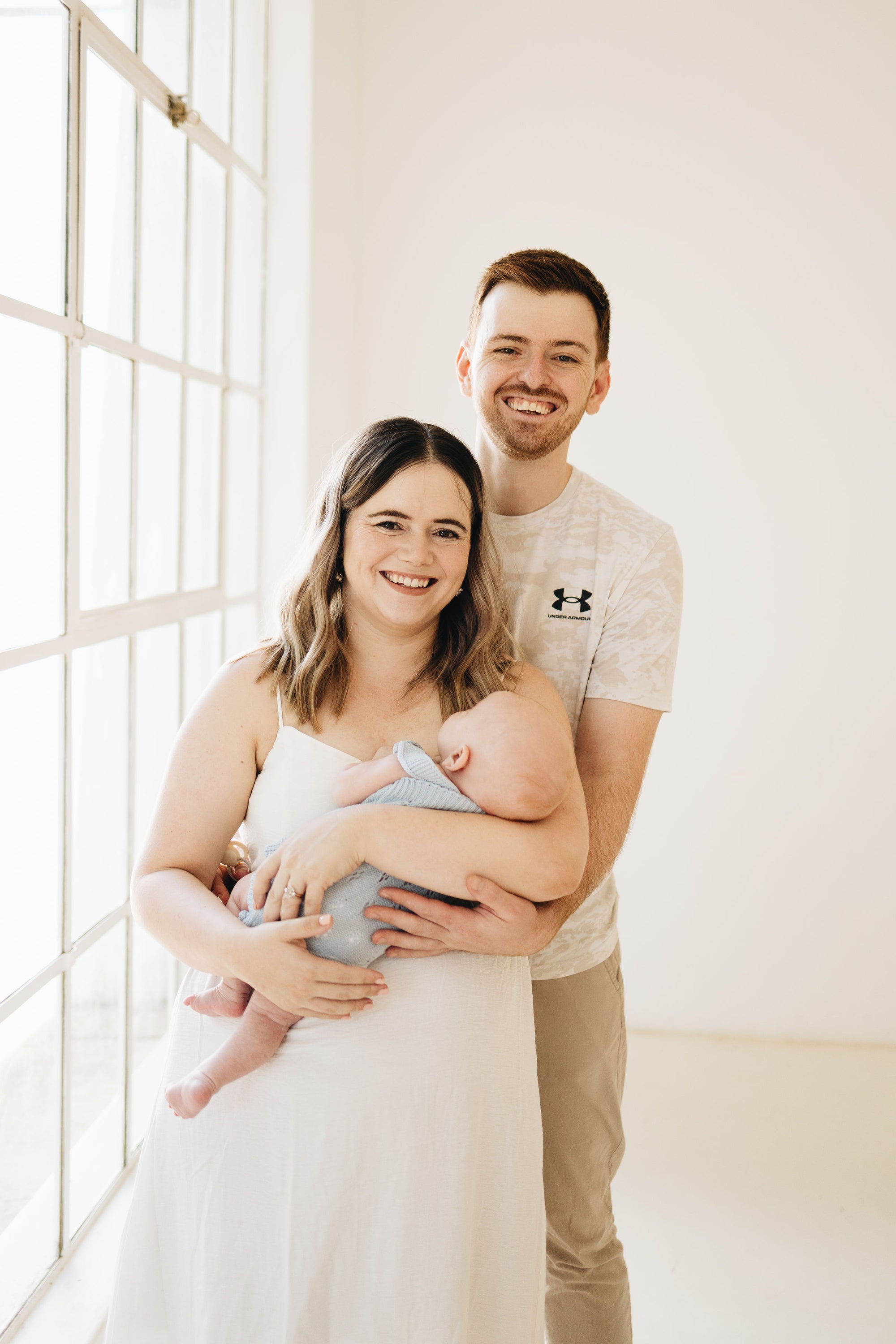 A smiling couple and their baby in a bright, minimalist room. The woman in a white dress holds a baby wrapped in a light blue blanket, while the man behind her wears a light gray Under Armour t-shirt and khaki pants.